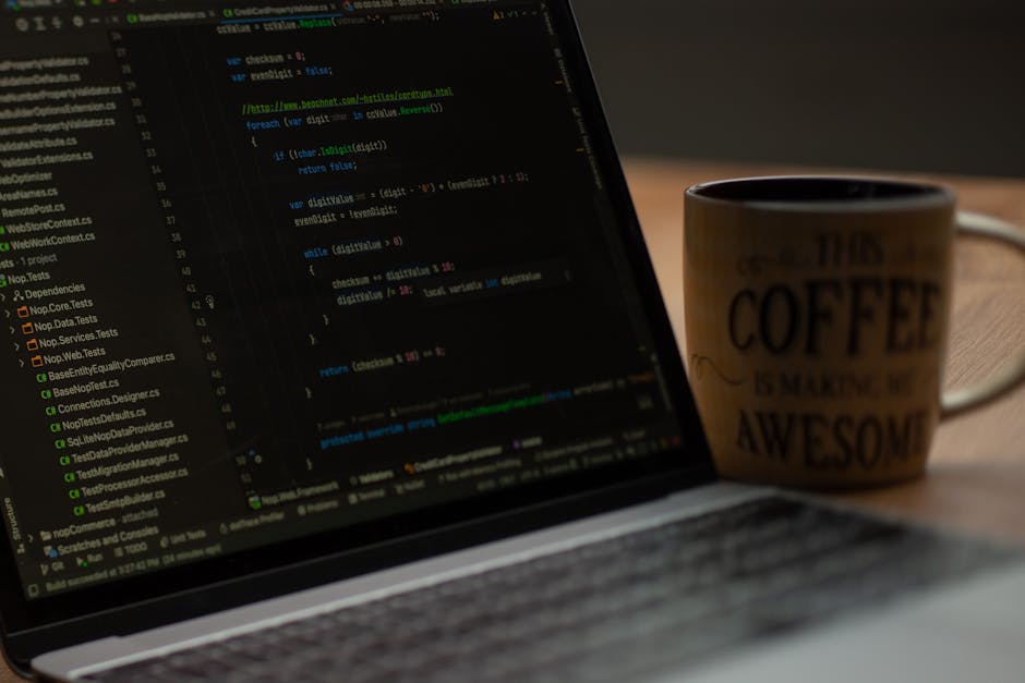 A close-up view of a laptop on a wooden table displaying lines of colorful coding text on its screen. Next to the laptop, there is a ceramic coffee mug with a beige background featuring black text tha